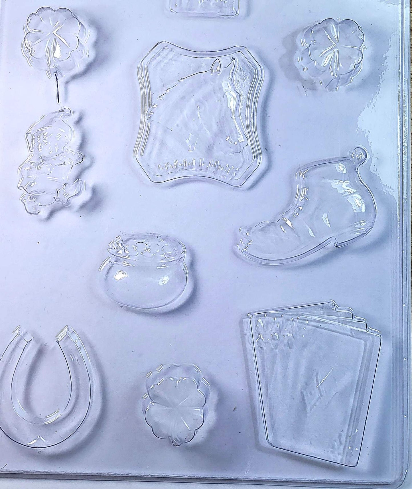 A clear plastic chocolate mould featuring shapes of a three-leaf clover, a horse shoe, a leprechaun, a pot of gold, a horse head with a good luck sign, a lucky boot, and four aces from playing cards.
