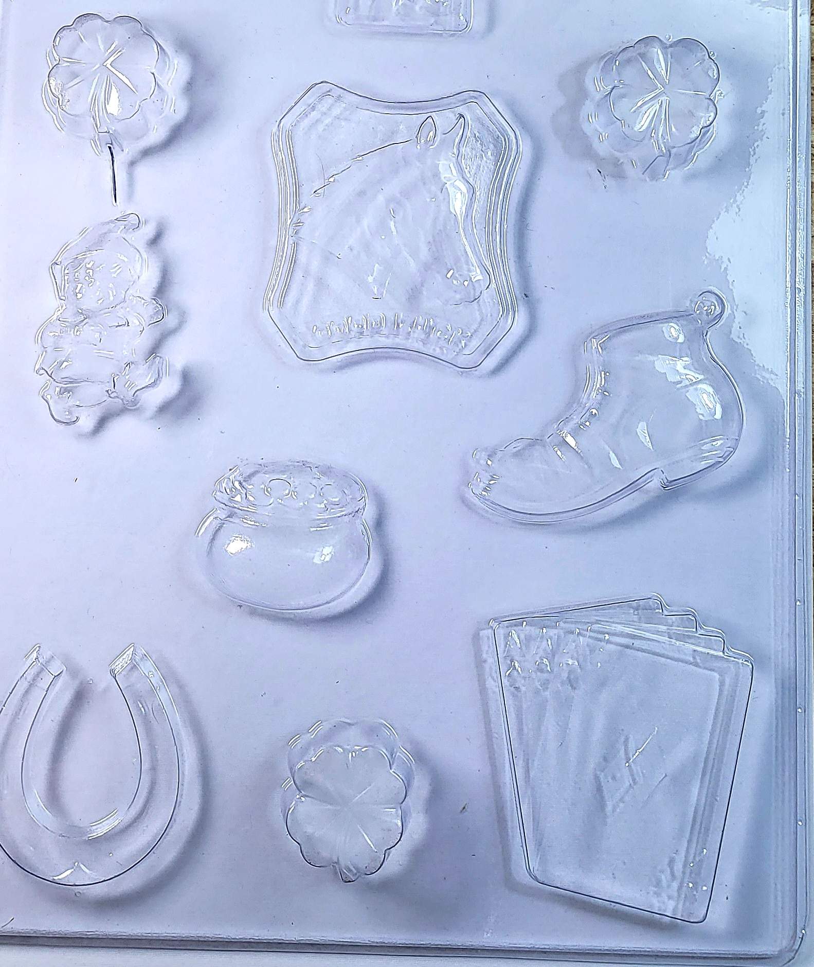 A clear plastic chocolate mould featuring shapes of a three-leaf clover, a horse shoe, a leprechaun, a pot of gold, a horse head with a good luck sign, a lucky boot, and four aces from playing cards.